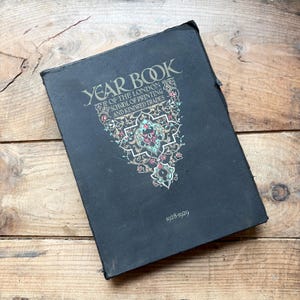 Pode incluir: Um livro de capa dura preta intitulado Year Book of the London School of Printing and Kindred Trades 1928-1929. A capa apresenta padrões decorativos de filigrana floral em dourado e turquesa abaixo do texto.