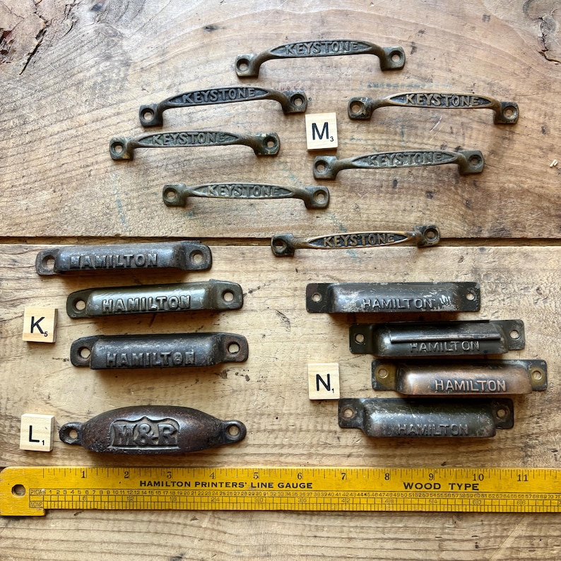 May include: A collection of vintage metal drawer pulls and bin handles featuring embossed text including Keystone, Hamilton, and M&R. The hardware is arranged in groups labeled with small wooden tiles K, L, M, and N, alongside a yellow 11-inch ruler.