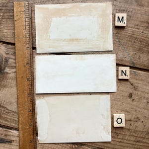 May include: Three rectangular, off-white vintage cards with aged edges are arranged on a wooden surface. A wooden ruler is positioned along the left side. Scrabble tiles with the letters M, N, and O are placed nearby.