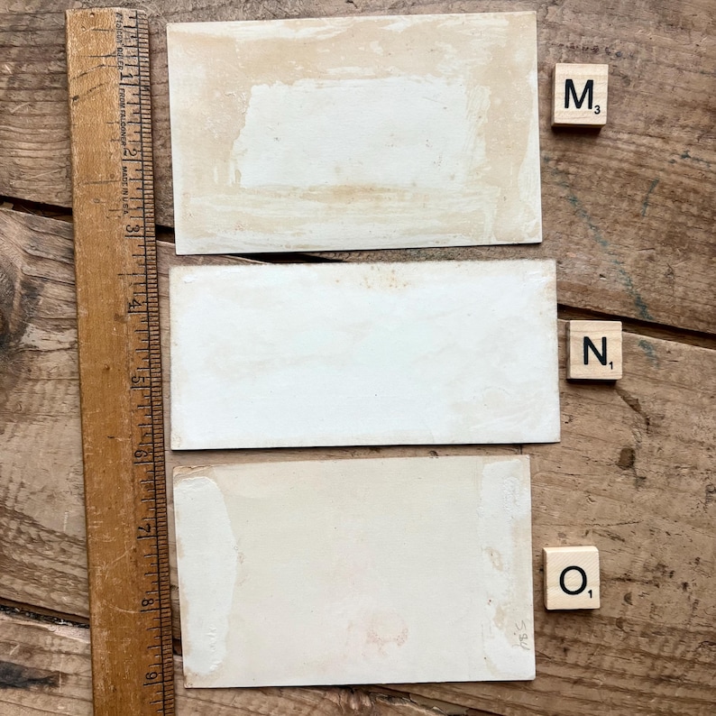 May include: Three rectangular, off-white vintage cards with aged edges are arranged on a wooden surface. A wooden ruler is positioned along the left side. Scrabble tiles with the letters M, N, and O are placed nearby.