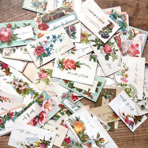 May include: A collection of vintage floral postcards with various designs. The cards feature roses, other flowers, and handwritten text in a variety of colors, including pink, red, and blue. The cards are scattered on a wooden surface.