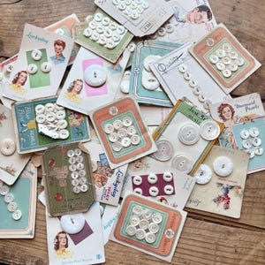 May include: A selection of vintage pearl buttons on original cards. The cards display illustrations of women and text such as "Luckyday" and "Superior Quality." The white buttons are of various sizes and arrangements.