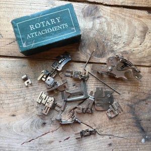 Puede incluir: Un conjunto de accesorios rotativos para máquinas de coser antiguas y una caja verde azulado con la inscripción "ROTARY ATTACHMENTS" en blanco. Los accesorios plateados están dispersos sobre una superficie de madera.