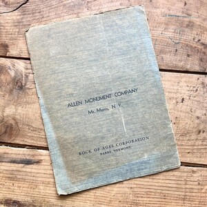 Puede incluir: Un documento de papel azul claro vintage con el texto "ALLEN MONUMENT COMPANY Mt. Morris, N.Y." y "ROCK OF AGES CORPORATION BARRE, VERMONT." El papel tiene un aspecto desgastado y est&aacute; sobre un fondo de madera.