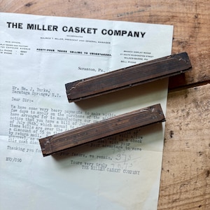 May include: Two rectangular, dark brown wooden printing blocks with engraved decorative borders, positioned diagonally across a vintage letterhead from The Miller Casket Company, which features black printed text on an off-white paper background.