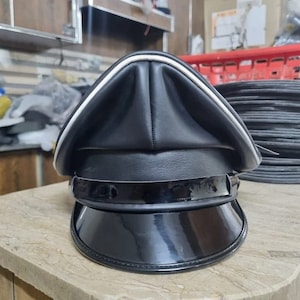 Puede incluir: Una gorra de pico de cuero negro con un borde blanco alrededor del borde. La gorra tiene un acabado negro brillante y un ala ancha.