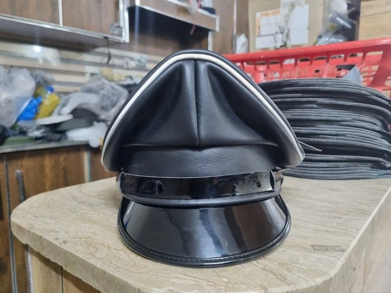 Puede incluir: Una gorra de pico de cuero negro con un borde blanco alrededor del borde. La gorra tiene un acabado negro brillante y un ala ancha.