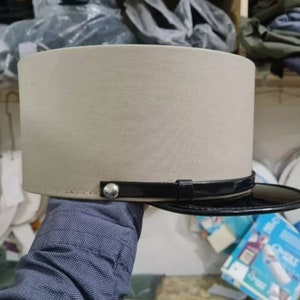 以下が含まれることがあります： ベージュとブラックの帽子で、ブラックのバンドとシルバーのボタンが付いています。帽子は柔らかな素材で作られており、わずかに丸みを帯びたクラウンを持っています。