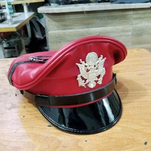 Könnte beinhalten: Ein roter Lederhut mit schwarzem Lacklederschirm und silbernem Emblem. Der Hut hat eine gewölbte Oberseite und ein schwarzes Band um den Sockel. Eine silberne Schnalle sichert einen schwarzen Riemen.