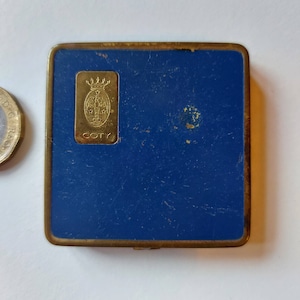 May include: A blue metal compact with a gold trim and a gold emblem featuring a crown and the word "COTY" on it.