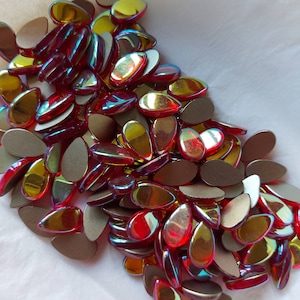 May include: A large pile of iridescent teardrop-shaped glass beads in shades of red and gold. The beads are scattered on a white surface.