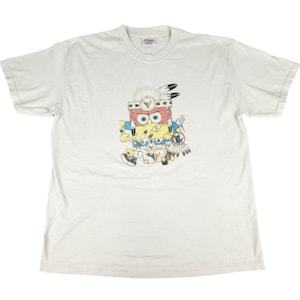 May include: White t-shirt featuring a cartoon character dressed in Native American attire. The character has a red and yellow face, wearing a feathered headdress and holding a dreamcatcher. The shirt is a crew neck.
