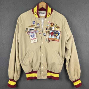Könnte beinhalten: Beige Bomberjacke mit einem bordeauxroten und gelb gestreiften Kragen, Bündchen und Saum. Die Jacke hat verschiedene Anstecknadeln und Aufnäher, darunter ein "Canon"-Sponsorenschild und einen Aufnäher für das Albuquerque International Balloon Fiesta 1985.