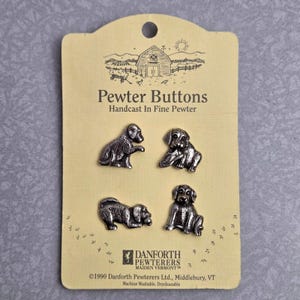 May include: Four silver-colored pewter buttons shaped like dogs, attached to a light yellow display card. The card features the text Pewter Buttons, Handcast In Fine Pewter, and Danforth Pewterers Maiden Vermont, with a 1990 copyright date.