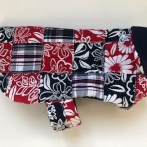 May include: A patchwork dog coat featuring a mix of floral and plaid patterns in red, white, and navy blue. The coat has a rounded shape and a sleeve for one leg. The coat is designed to keep a dog warm.