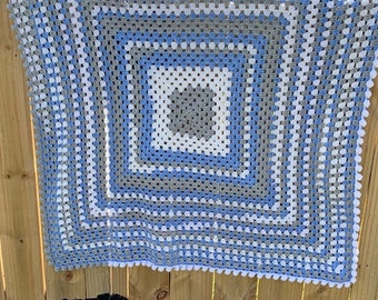 May include: A crocheted baby blanket in shades of blue, gray, and white with a white pom-pom trim.