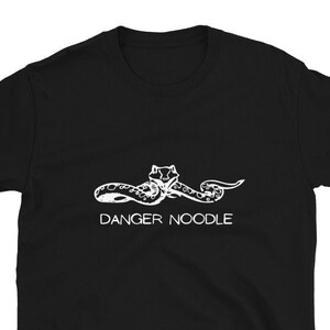 May include: Black t-shirt featuring a white graphic of a snake with the text "DANGER NOODLE" below it. The snake is stylized with a detailed head and a curved body. The shirt is made of a soft material.