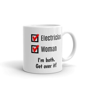 May include: White ceramic mug with a black and red design. The mug has a checklist with two boxes checked. The boxes are labeled "Electrician" and "Woman". The text below the checklist reads "I'm both. Get over it!"