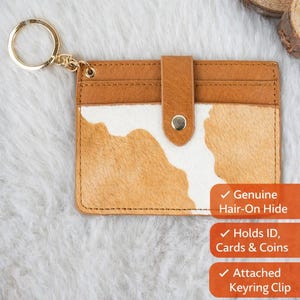 May include: Brown and white cowhide keychain wallet with a gold keyring. The wallet features a brown leather flap with a snap closure. The wallet is made of genuine hair-on hide and holds ID, cards, and coins. The text reads "Carry Your Essentials in Western Style!"