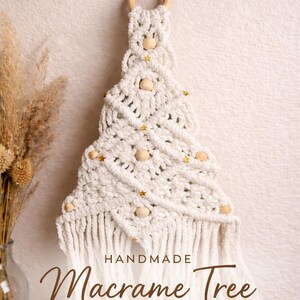 May include: Handmade macrame Christmas tree wall hanging. The off-white tree features wooden beads and gold star accents. The tree is attached to a wooden ring and a decorative metal hook.