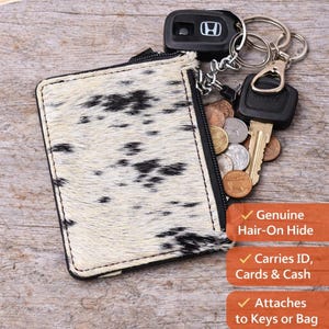 May include: Black and white cowhide keychain wallet with a zipper, holding keys, coins, and a car key. The wallet is made of genuine hair-on hide and carries ID, cards, and cash. It attaches to keys or a bag.