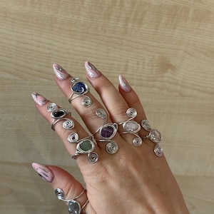 May include: A collection of seven silver-toned wire-wrapped rings, each featuring a unique polished gemstone. The stones include blue, black, purple, green, and clear varieties, all secured by intricate metal wire spirals and loops.