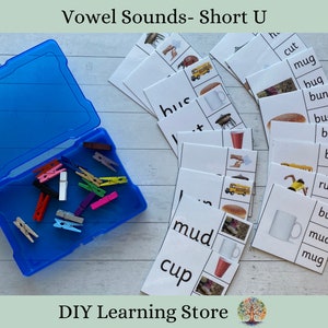 May include: A set of 12 flash cards with pictures and words for learning the short "u" vowel sound. The cards feature words like "bus", "cut", "mug", "bug", "bun", "mud", and "cup". The cards are laid out on a white wooden surface with a blue container of colorful clothespins.