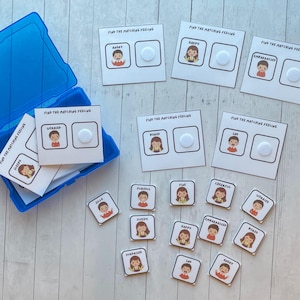 May include: A set of 12 colorful cards with illustrations of people expressing different emotions. Each card has the word "FIND THE MATCHING FEELING" and the name of the emotion. The cards are arranged on a white wooden surface.