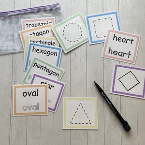 May include: A set of colorful flashcards with various shapes and their names, including trapezoid, octagon, rectangle, hexagon, pentagon, oval, heart, and square. The cards are laid out on a wooden surface with a black marker.