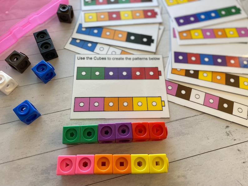 May include: A set of colourful plastic cubes with different shapes on each side. The cubes are arranged in a line, with the shapes facing up. The text "Use the Cubes to create the patterns below" is printed on a white card.