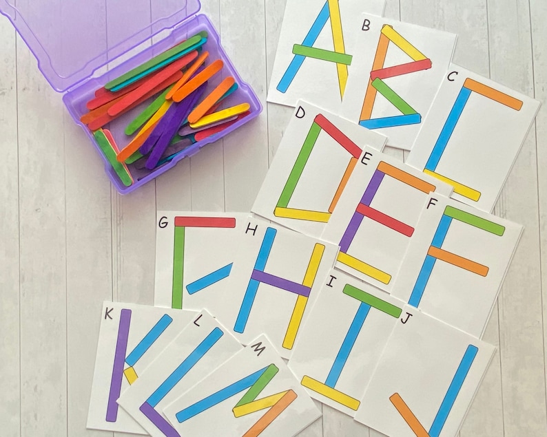May include: A set of alphabet letter cards with the letters A through M printed on them. Each card has a different letter and a corresponding design made of colorful popsicle sticks. The cards are on a white wooden surface.