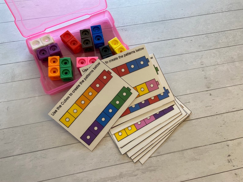May include: A set of colourful plastic cubes in a pink container with cards that have instructions to create patterns using the cubes. The cards show different colour patterns with white circles.