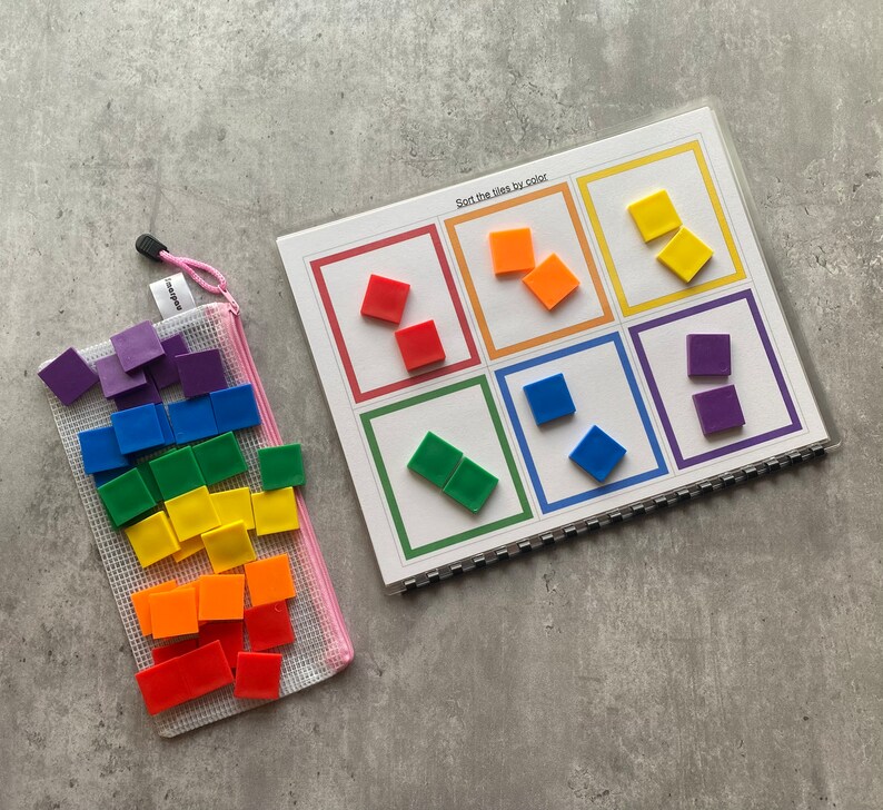 May include: A color sorting activity set featuring a workbook with color-coded squares and a mesh bag filled with matching colored tiles. The workbook has the text "Sort the tiles by color". The tiles are red, orange, yellow, green, blue, and purple.