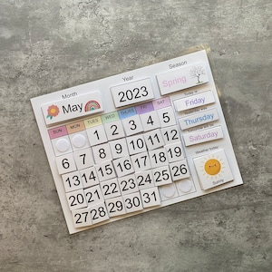 Pu&ograve; includere: Un calendario magnetico colorato per maggio 2023 con una grafica di fiori, un arcobaleno e un'icona del sole. Il calendario include i giorni della settimana, i numeri e una sezione per la stagione, il tempo e il giorno della settimana.