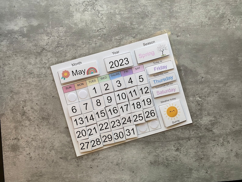 Pu&ograve; includere: Un calendario magnetico colorato per maggio 2023 con una grafica di fiori, un arcobaleno e un'icona del sole. Il calendario include i giorni della settimana, i numeri e una sezione per la stagione, il tempo e il giorno della settimana.