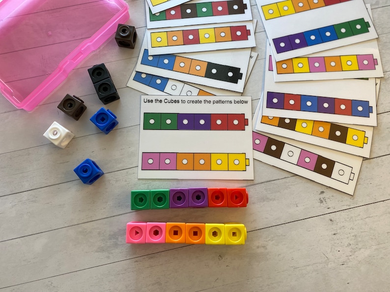 May include: A set of colourful plastic cubes with different shapes on each side. The cubes are arranged in a line to create a pattern. The text "Use the Cubes to create the patterns below" is printed on a white card.