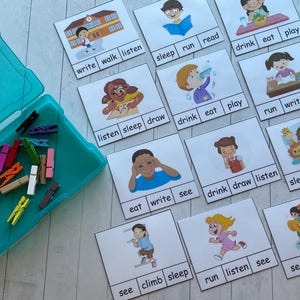 May include: A set of 12 colorful picture cards with various images of children performing different actions. Each card has four words printed on it, with one word per row. The words are: write, walk, listen, sleep, run, read, drink, eat, play, listen, sleep, draw, drink, eat, play, run, write, walk, eat, write, see, drink, draw, listen, sleep, eat, play, see, climb, sleep, run, listen, see, see, play, walk.