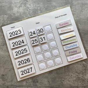 Pu&ograve; includere: Bacheca di calendario bianca con numeri neri e cerchi bianchi. La bacheca ha una sezione per gli anni, i giorni e i giorni della settimana. Gli anni sono 2023, 2024, 2025, 2026 e 2027. I giorni sono 24, 25, 30 e 31. I giorni della settimana sono domenica, luned&igrave;, marted&igrave;, mercoled&igrave;, gioved&igrave;, venerd&igrave; e sabato.