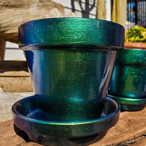 May include: A green and blue iridescent ceramic planter with a matching saucer. The planter has a smooth, shiny finish and a slightly textured surface.