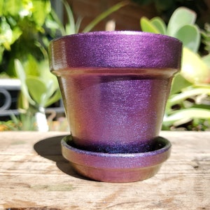 May include: A purple iridescent ceramic planter with a matching saucer. The planter has a textured surface and a slightly flared lip.