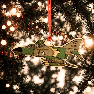 May include: A wooden Christmas ornament shaped like a military jet, painted in shades of green and tan with a red ribbon for hanging. The ornament features the text "FP 680" on the tail and a star emblem. The ornament is hanging on a Christmas tree.