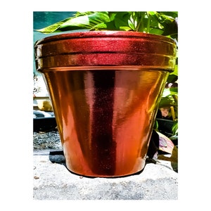May include: A large, metallic copper-colored flower pot with a wide rim. The pot has a glossy finish and is set against a backdrop of green foliage. The pot is designed for planting flowers or other plants.