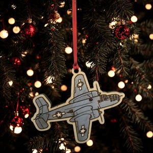 May include: Wooden Christmas ornament shaped like a vintage airplane, painted blue with white star accents. The ornament hangs from a red ribbon against a backdrop of a Christmas tree with red ornaments and twinkling lights.