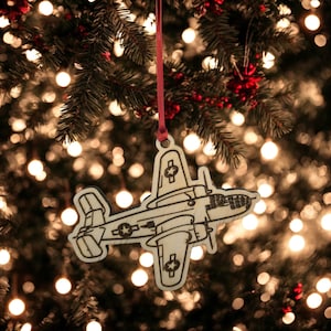 May include: A wooden airplane ornament with a red ribbon, hanging on a Christmas tree. The ornament is a light tan color with black etched details. The background is blurred with Christmas lights.
