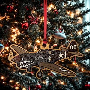 May include: Wooden airplane Christmas ornament with a red ribbon. The ornament is brown and black with a shark mouth design and the text "The Jacky C". The plane has a white star and the number "00" on the tail.