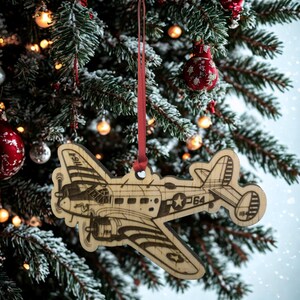 May include: Wooden airplane Christmas ornament with a red ribbon for hanging. The ornament features an engraved design of a vintage airplane with the number "1164" and a star. The ornament is hanging on a Christmas tree.