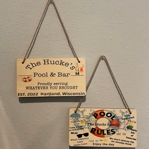 May include: Two wooden signs with a pool theme. The top sign reads "The Hucke's Pool & Bar" and "Proudly serving WHATEVER YOU BROUGHT EST. 2022 Hartland, Wisconsin". The bottom sign reads "POOL RULES The Hucke Family" with a life preserver graphic and other pool-related graphics. The sign also includes the text "Stay hydrated", "Have fun", "Apply lots of sunscreen", "We don't skinny dip we chunky dunk", "Soak up the", and "Enjoy the day".