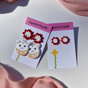 Puede incluir: Dos pares de pendientes en tarjetas blancas con el texto "SapphicStopAU". Un par presenta pendientes en forma de flor roja y blanca con una cara de animal de dibujos animados blanca colgando de cada uno. El otro par presenta pendientes en forma de flor roja y blanca.