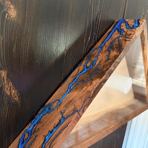 May include: A triangular wooden shelf featuring a natural wood grain finish and a striking electric blue resin inlay that mimics the branching pattern of a lightning strike. The shelf includes a small white cylindrical dowel for storage.