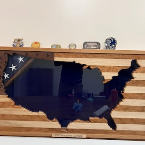 May include: A wooden frame with a cutout of the United States of America, with a blue background. The frame is decorated with an American flag design and has a plaque at the bottom that reads "CPT Angeline Lienhop".